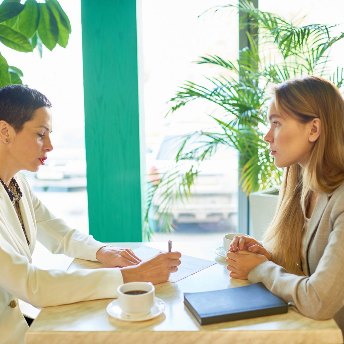 Side view portrait of two modern young women discussing work sitting at table in cafe during business meeting or job interview